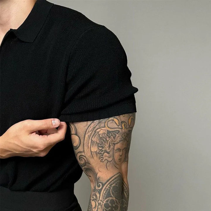 A person in a fitted black quarter zip polo with ribbed detailing rolls up their sleeve, revealing a detailed tattoo on their upper arm featuring a classical warrior helmet and shield design.