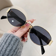 A hand with manicured nails holds a pair of vintage-inspired sunglasses with black lenses and thin gold metal frames. The person is wearing a gray knit sweater, showcasing effortless Italian style. The background is out of focus.