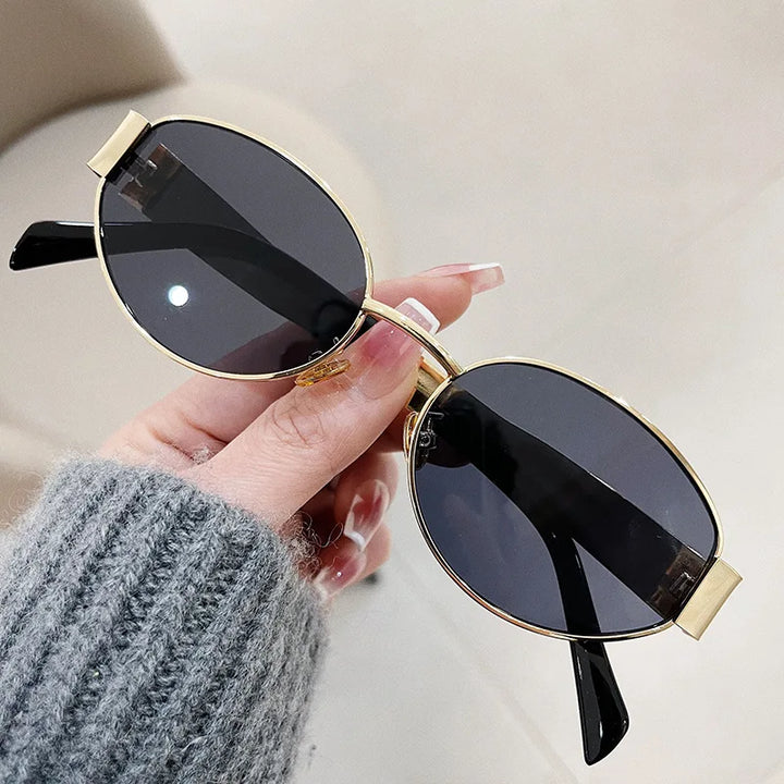 A hand with manicured nails holds a pair of vintage-inspired sunglasses with black lenses and thin gold metal frames. The person is wearing a gray knit sweater, showcasing effortless Italian style. The background is out of focus.