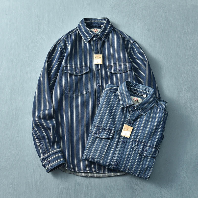 Two blue denim shirts with white vertical stripes, featuring a relaxed fit and buttoned collars, are neatly arranged on a plain blue surface. Both have chest pockets and tan tags attached to the button plackets.