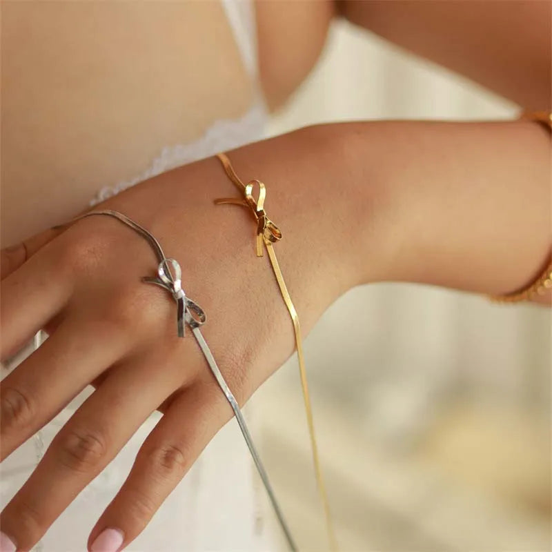 A close-up of a womans hand and wrist wearing two delicate ribbon-shaped bracelets, one gold and one silver, showcases elegant minimalist jewelry against a soft, neutral background.