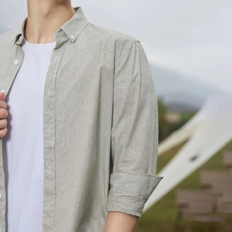 A person wearing a classic pinstripe shirt over a white T-shirt stands outdoors with rolled-up sleeves. The light gray cotton button-up is stylish and comfortable, set against a blurred background of greenery and distant structures.