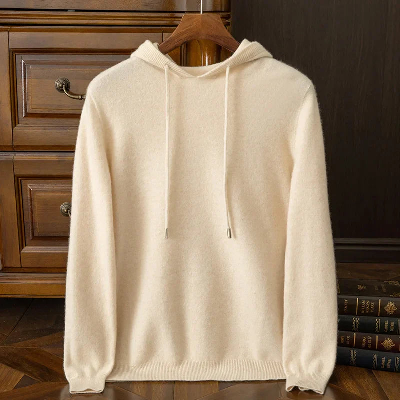 A cream-colored, long-sleeve cashmere blend hooded sweater with drawstrings hangs on a wooden hanger, positioned in front of a wooden dresser and beside a stack of books.
