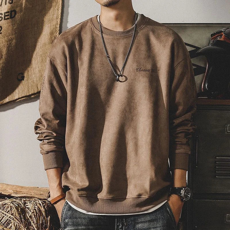 A man wears a relaxed fit crewneck in a loose brown, earth-toned style with a round neckline, black necklace with a ring pendant, and watch. His face isn’t visible as he stands casually with hands in his pockets.