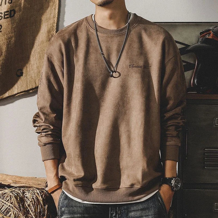 A man wears a relaxed fit crewneck in a loose brown, earth-toned style with a round neckline, black necklace with a ring pendant, and watch. His face isn’t visible as he stands casually with hands in his pockets.