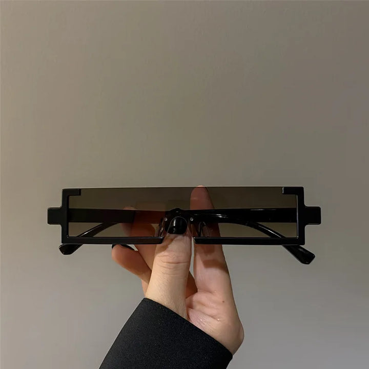 A hand is holding a pair of rectangular, geometric sunglasses with thick black frames and dark lenses that offer UV400 protection against a plain gray background.