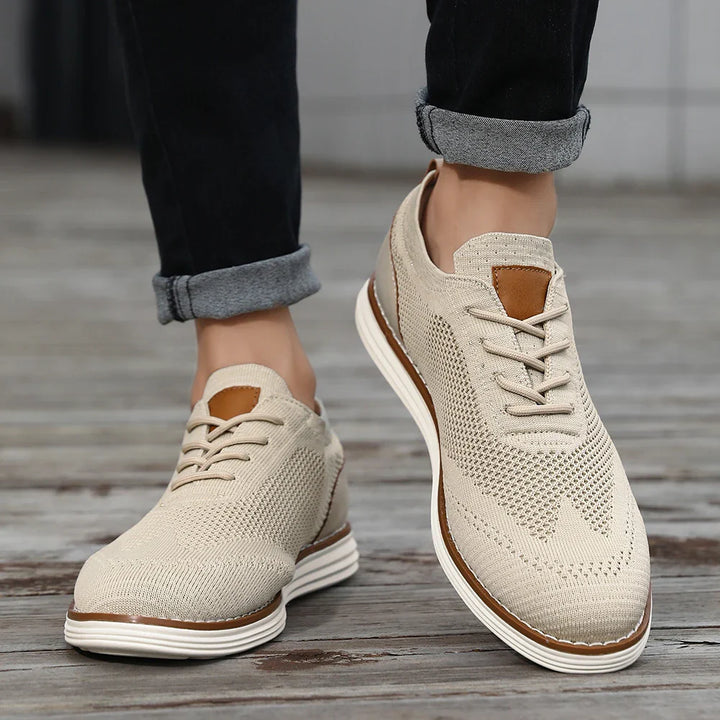 A person wearing breathable knit sneakers with white soles and brown accents stands on a wooden surface, jeans cuffed above the ankles, showcasing a relaxed casual style.