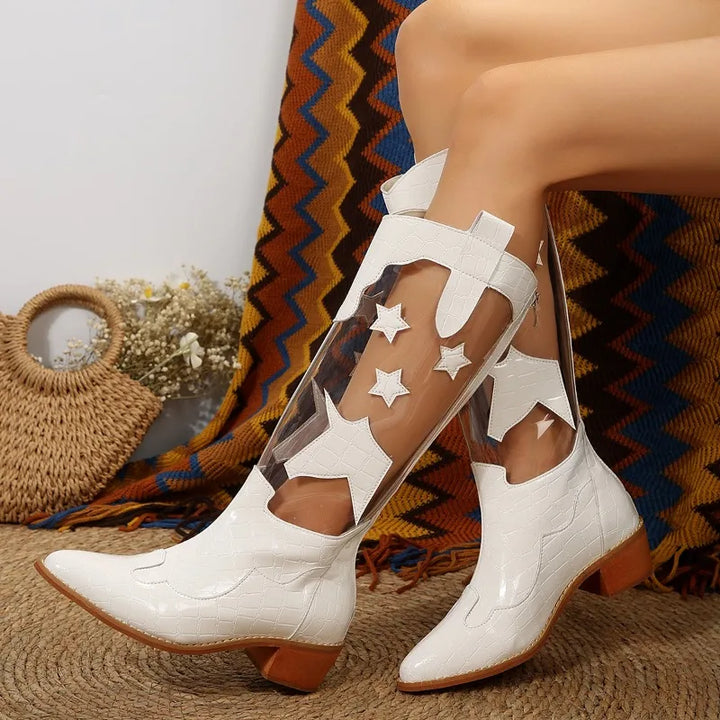 A person wearing knee-high white clear panel boots with star-shaped cutouts sits against a patterned blanket, with a woven bag and dried flowers in the background.