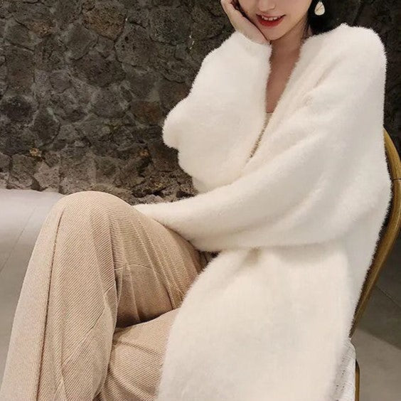 A woman sits on a chair in a long, fluffy white cardigan with a soft silken feel and beige pants, one arm resting on her knee and the other touching her face. A stone wall is visible in the background.