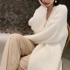 A woman sits on a chair in a long, fluffy white cardigan with a soft silken feel and beige pants, one arm resting on her knee and the other touching her face. A stone wall is visible in the background.