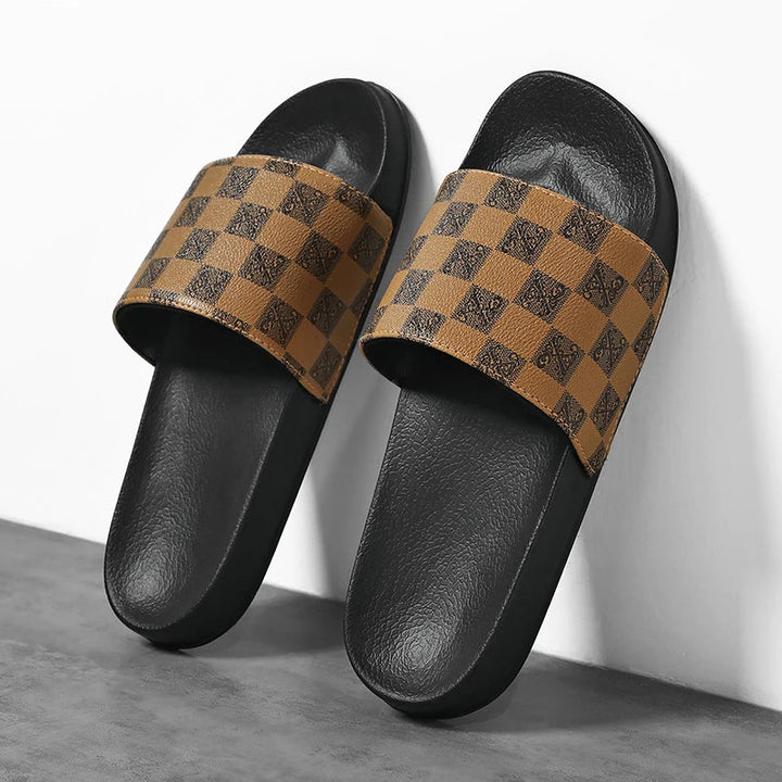 A pair of black streetwear slides with brown straps featuring a checkered pattern, positioned sideways against a white wall and gray floor.
