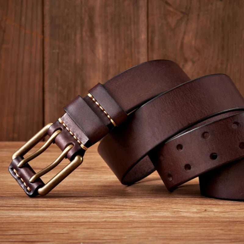 A dark brown cowhide leather belt with a brass buckle is coiled on a wooden surface with a wooden background. The durable belt features visible stitching near the buckle and several holes along its length.