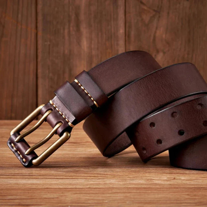 A dark brown cowhide leather belt with a brass buckle is coiled on a wooden surface with a wooden background. The durable belt features visible stitching near the buckle and several holes along its length.