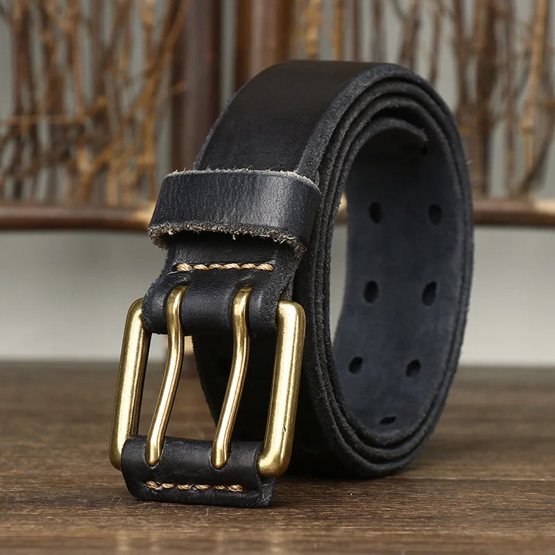 A rolled-up black genuine leather belt with a double-prong copper buckle and visible stitching rests on a wooden surface, hinting at heritage craftsmanship against a blurred background of wooden sticks.