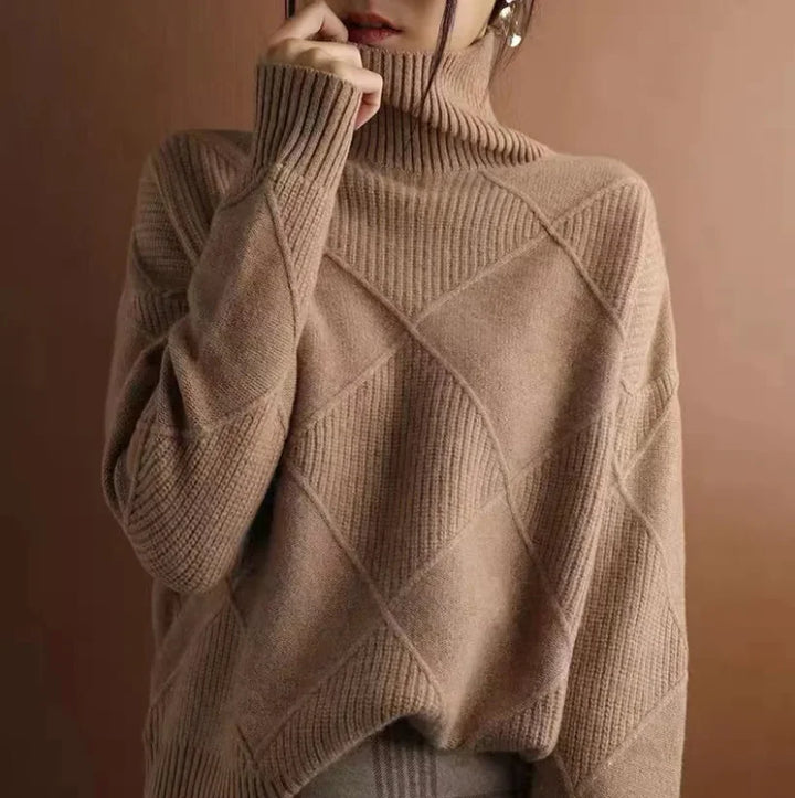 A person wearing a cozy, loose-fitting, diamond knit cashmere turtleneck sweater stands against a matching brown background. The face is partially obscured by the sweater collar and their hand.