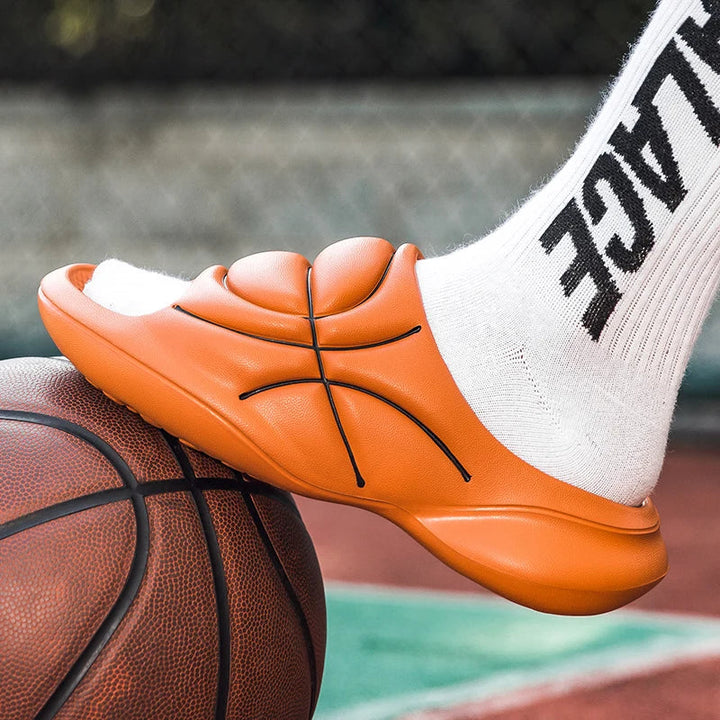 A person wearing white socks and orange basketball slip ons, featuring a cushioned footbed and designed to look like a basketball, rests their foot on an actual basketball on an outdoor court.