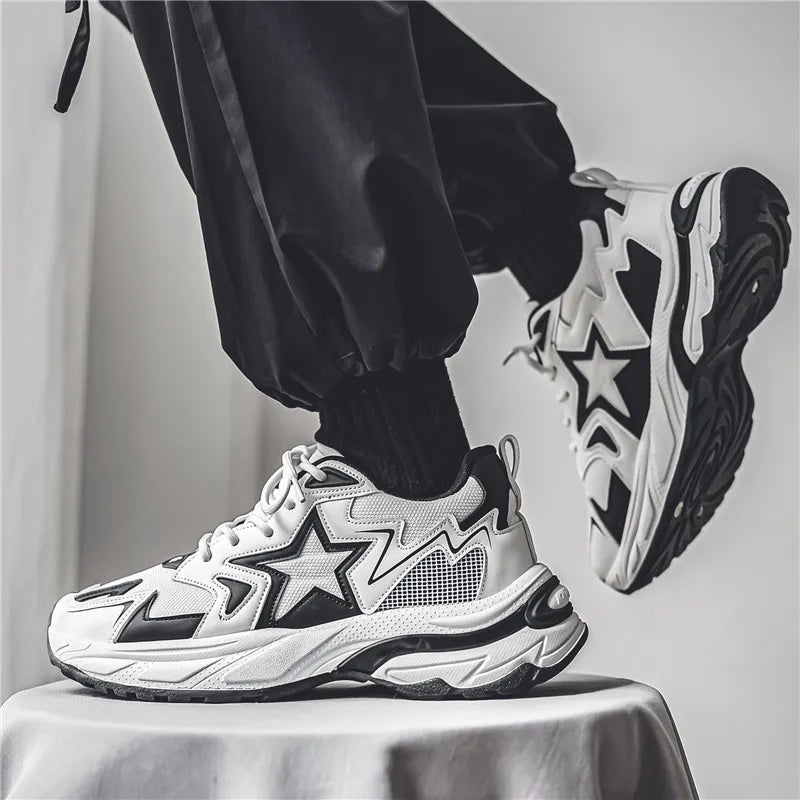 A person wearing black pants and black socks stands on a stool, showcasing chunky white sneakers with bold black star designs, a breathable mesh upper, and striking patterns on the sides and soles.