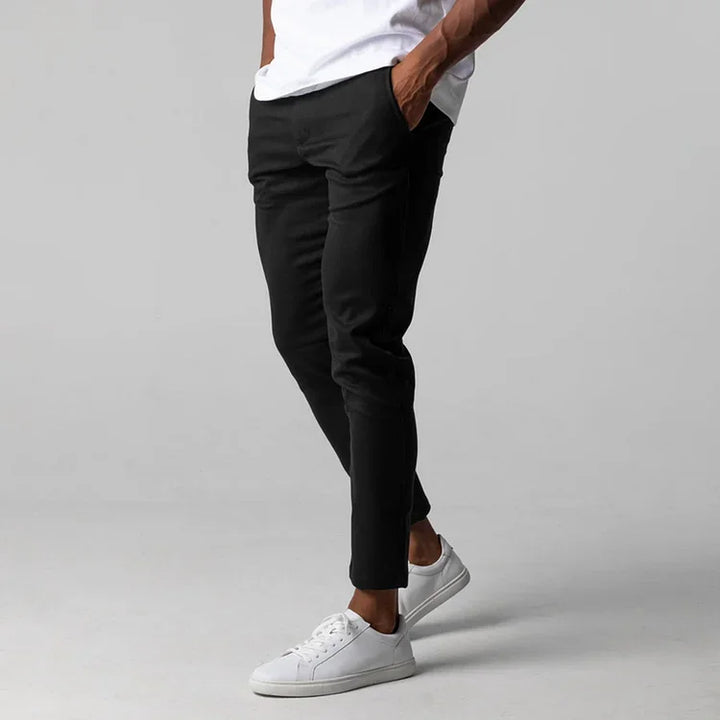 A person stands with hands in pockets, wearing black slim fit ankle length pants, a white T-shirt, and white sneakers against a plain gray background. Their face is not visible.
