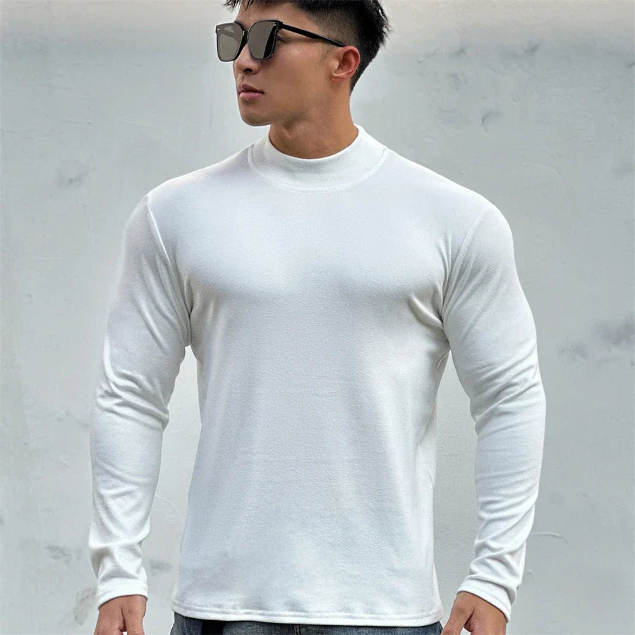 A man wearing black sunglasses and a fitted, long-sleeve white shirt with a mock neck collar stands against a light gray wall, looking to the side with a confident stance.