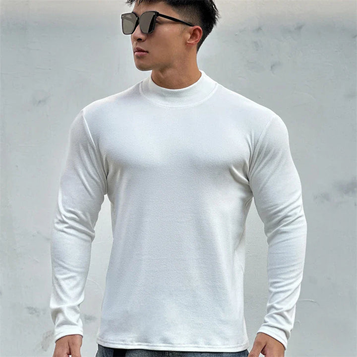 A man wearing black sunglasses and a fitted, long-sleeve white shirt with a mock neck collar stands against a light gray wall, looking to the side with a confident stance.