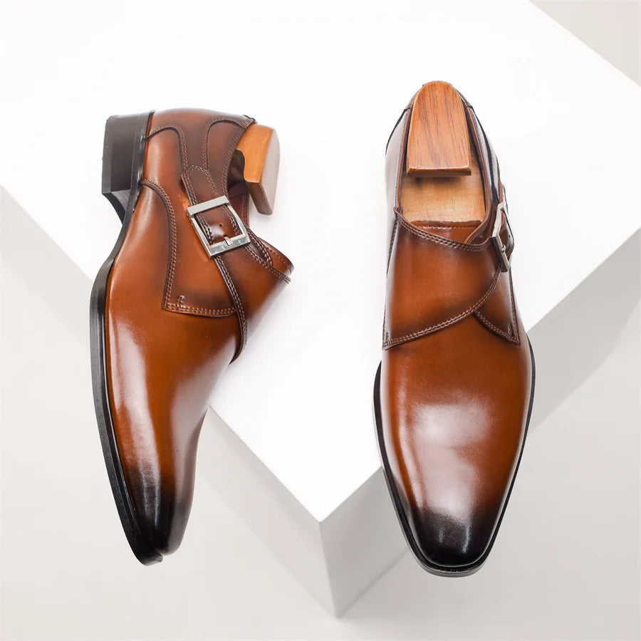 A pair of polished brown Italian leather monk strap shoes, each with a silver buckle, displayed on a white geometric surface. One handcrafted shoe is upright with a wooden shoe tree, the other lies flat.