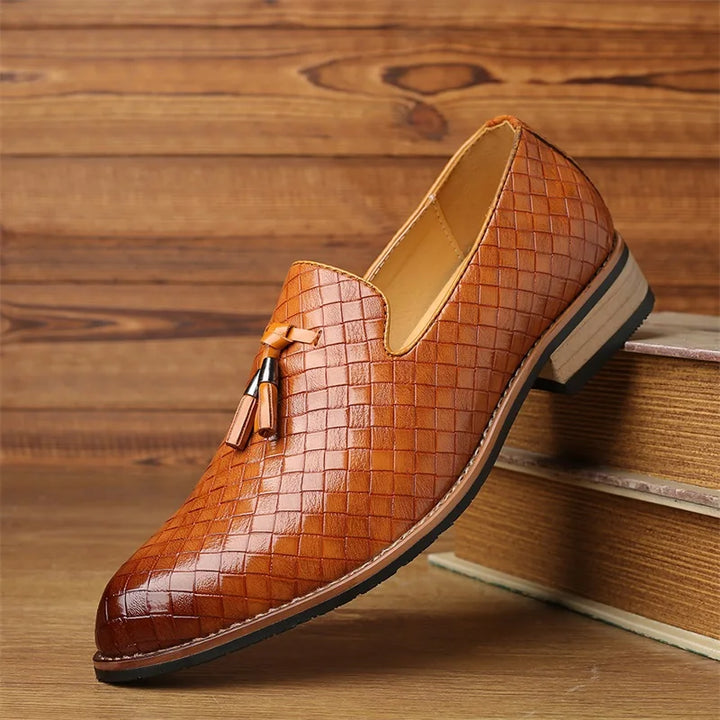 A single brown basketweave pattern loafer with a tassel detail, crafted from real leather shoes, displayed on top of stacked books against a wooden background.