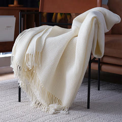 A cream-colored knit waffle blanket with fringed edges is draped over a black metal chair in a cozy, carpeted room with brown furniture, adding a stylish boho decorative accent to the space.