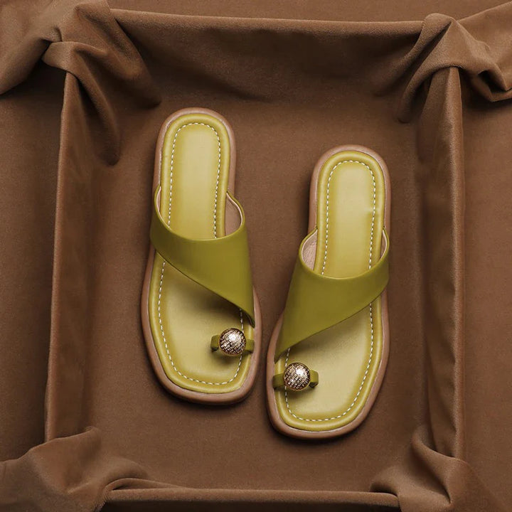 A pair of green, square-toe slides with an asymmetrical strap and a decorative gold sphere on the toe, displayed on brown fabric—perfect for those seeking fashionable slides with modern flair.