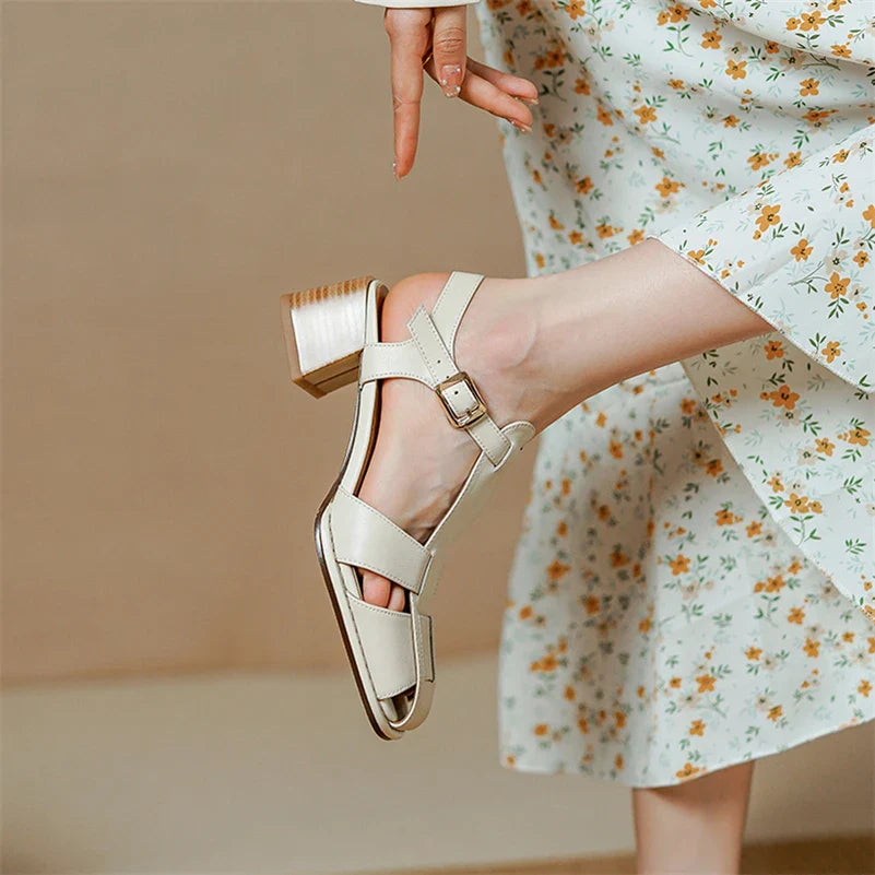 A person wearing a floral dress shows off a beige heeled sandal with ergonomic arch support and a buckle strap, lifting one foot slightly against a neutral background.
