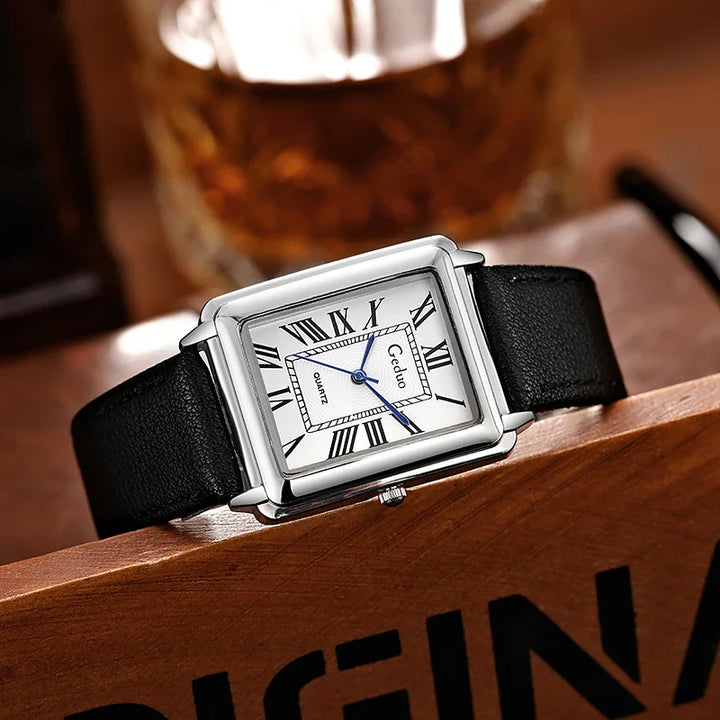 A rectangular luxury watch with a white dial, Roman numerals, blue hands, and a premium leather band rests on a wooden surface, with a blurry glass in the background.