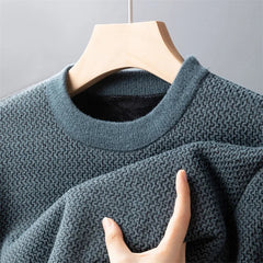 A close-up of a textured, dark green high-quality cotton sweater hanging on a wooden hanger, with a hand gently holding part of the fabric to show its softness and knit detail.
