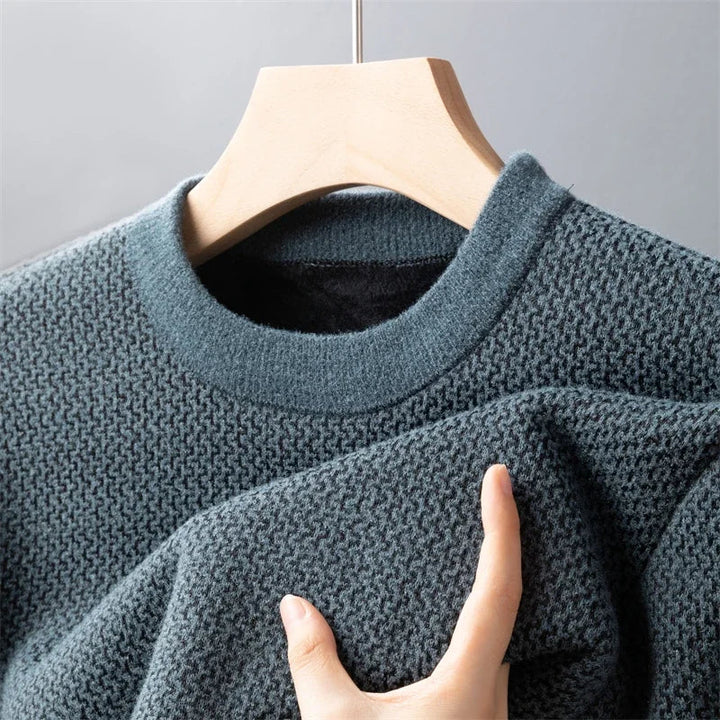 A close-up of a textured, dark green high-quality cotton sweater hanging on a wooden hanger, with a hand gently holding part of the fabric to show its softness and knit detail.
