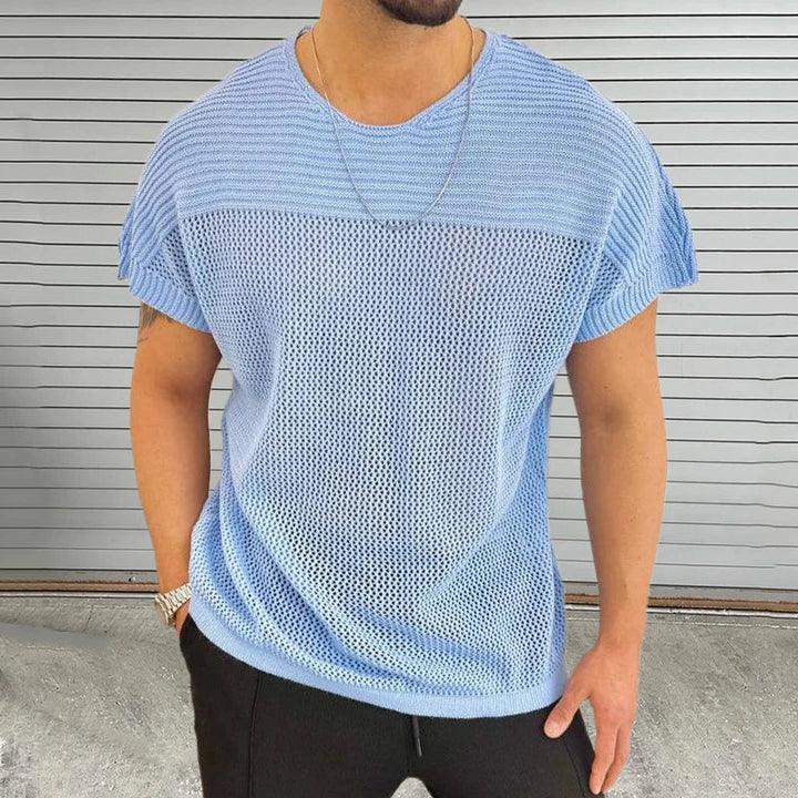 A man in a light blue mesh knit tee with a breathable design and black pants stands before a gray metal garage door. He accessorizes his streetwear look with a chain necklace and watch. His face is not visible.
