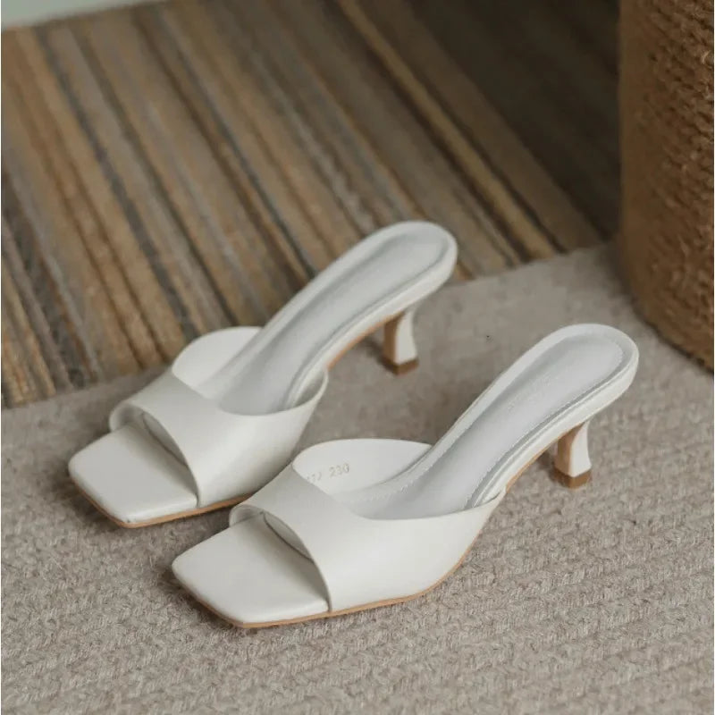 A pair of white heeled sandals with square open toes, ergonomic arch support, and cushioned insoles, placed on a beige carpet with a striped rug nearby.