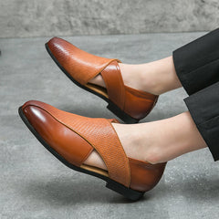 A person wearing stylish low profile loafers in two-tone brown genuine leather with a textured strap design and black trousers, sitting on a gray stone floor.