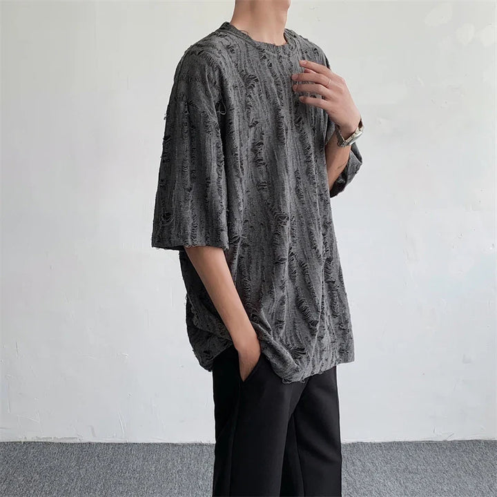 A person wearing an oversized, textured gray streetwear tee and black pants stands against a plain light-colored wall, with one hand in their pocket and the other touching their collarbone.