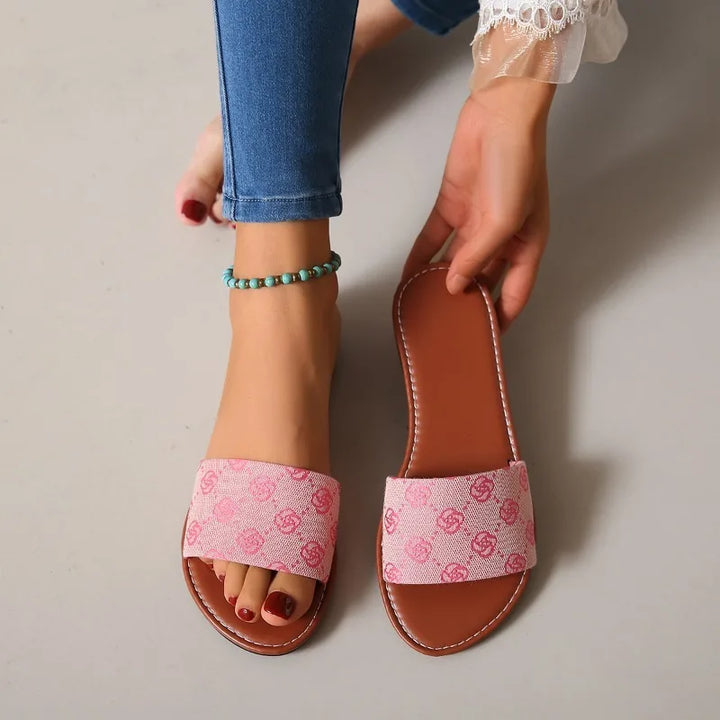 A person wearing blue jeans and an anklet tries on a pink open-toe slide silhouette sandal with a patterned strap; their toenails are painted red. Another timeless French Sole sandal is held in their hand.