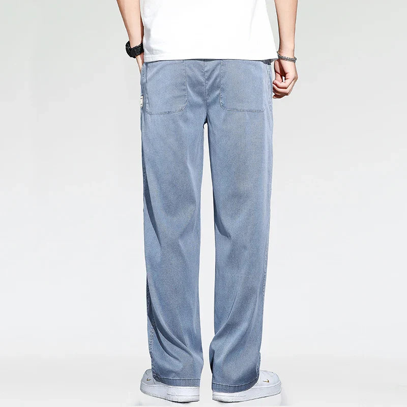 A person wearing summer loose men jeans, a white T-shirt, white sneakers, and a wristwatch is standing with their back to the camera against a plain light background.