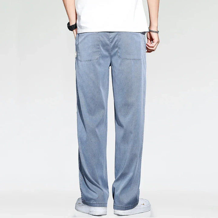 A person wearing summer loose men jeans, a white T-shirt, white sneakers, and a wristwatch is standing with their back to the camera against a plain light background.