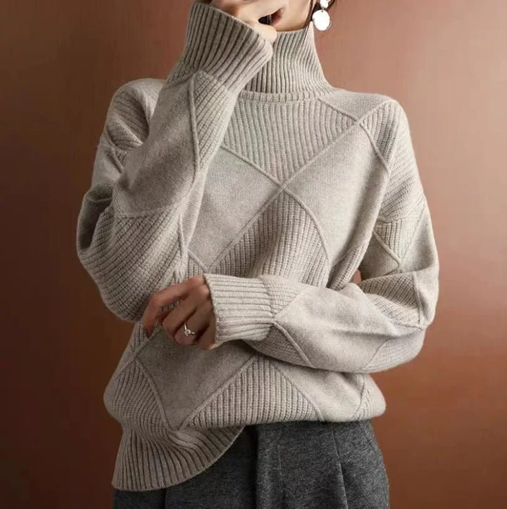 Person wearing a light beige, diamond knit turtleneck sweater with ribbed detailing and dark gray pants, standing against a plain brown background. The persons face is partially obscured by their hand.