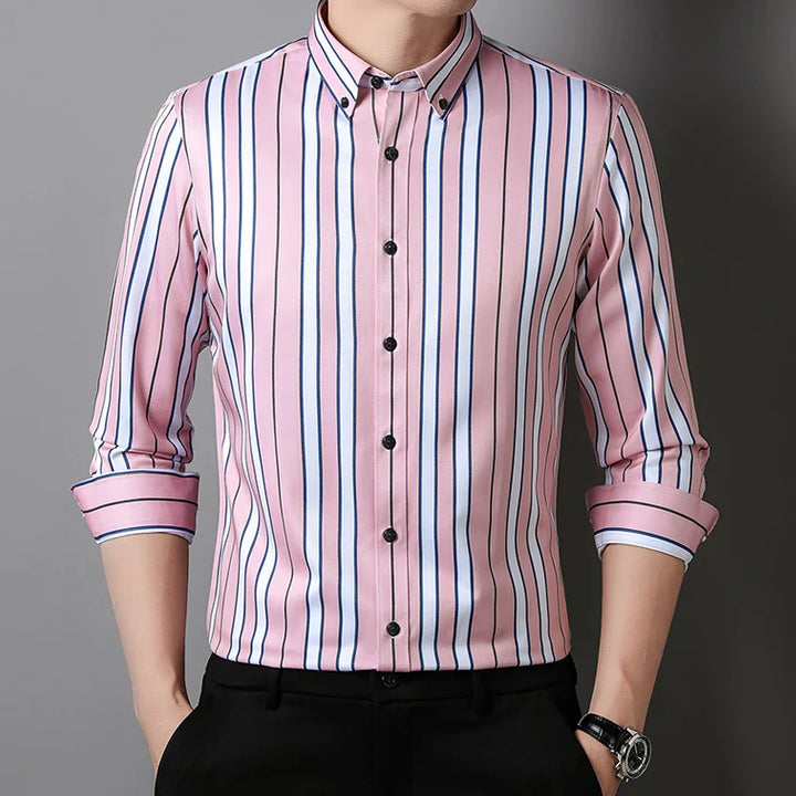 A person wearing a Hamilton Executive Classic Fit Shirt—a long-sleeved, button-up pink shirt with vertical blue, white, and black stripes. The sleeves are rolled up and hands are in pockets, showcasing this premium fabric shirt.
