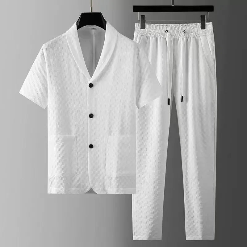 A white two-piece set features a short-sleeve button-up shirt with a textured pattern and two front pockets, paired with matching white pants with an elastic drawstring waistband, displayed on black hangers against a gray background.