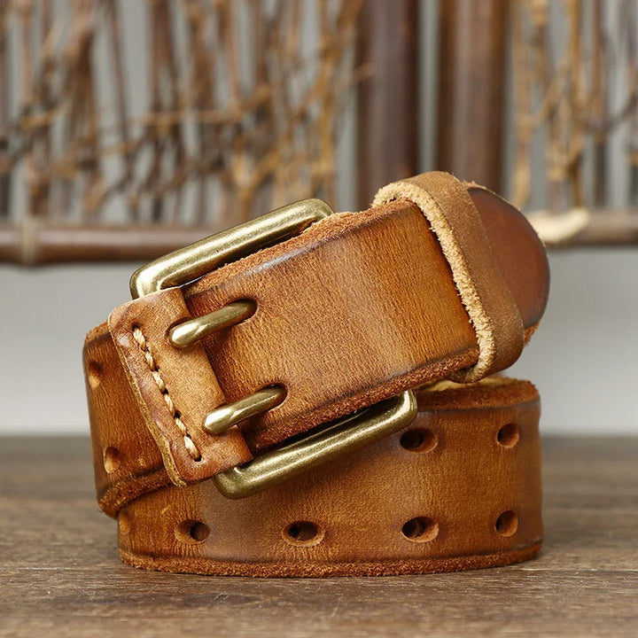 A coiled brown genuine leather belt with visible stitching and a copper buckle, featuring multiple oval holes, displayed on a wooden surface with a blurred rustic background that highlights heritage craftsmanship.