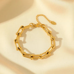 A gold statement chain bracelet with cylindrical links and small round accents, crafted from 316L stainless steel. Displayed on soft cream fabric, it features an adjustable extension chain and a small round tag.