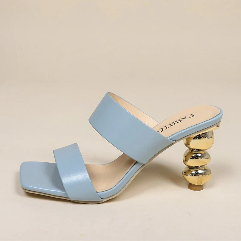 A stylish light blue sandal crafted from premium leather, featuring two wide straps and a unique gold sculpted heel shaped like stacked spheres. The square toe and modern, minimalist design complete the look.