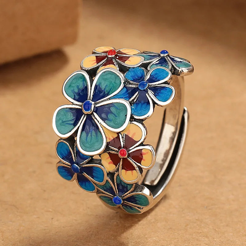 A silver ring decorated with vibrant colors in enamel flower designs—blue, yellow, and red—arranged closely together. The background is a warm brown surface.