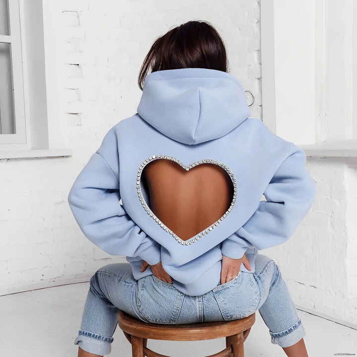 A person sits on a stool facing away, wearing a light blue heart-shaped open back hoodie with rhinestone trim, paired with light blue jeans in a bright, white room.