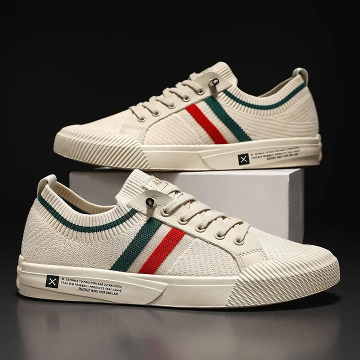 Two beige knit sneakers with a breathable mesh upper and white soles, featuring green and red side stripes, white laces, and a small text label on the outer sole. One shoe is elevated on a white box against a black background.
