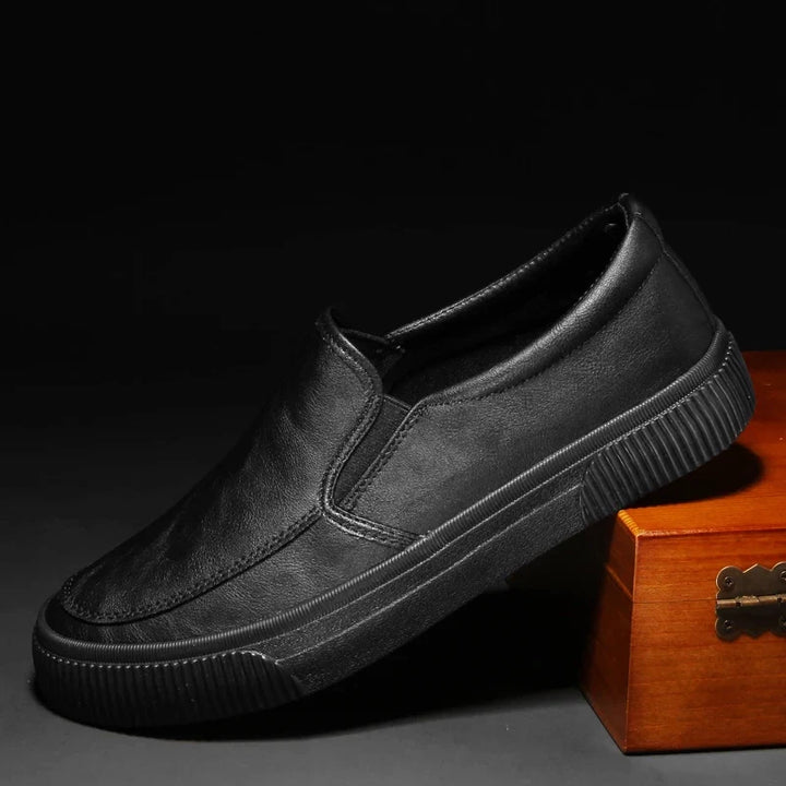A men’s slip-on black casual shoe with a hybrid rubber sole is displayed at an angle, resting one end on a wooden box against a dark background.
