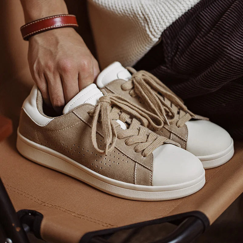 A person’s hand holds a beige Lowland Odyssey Sneaker with white accents and laces, next to another matching minimalist sneaker, both placed on a brown fabric surface.